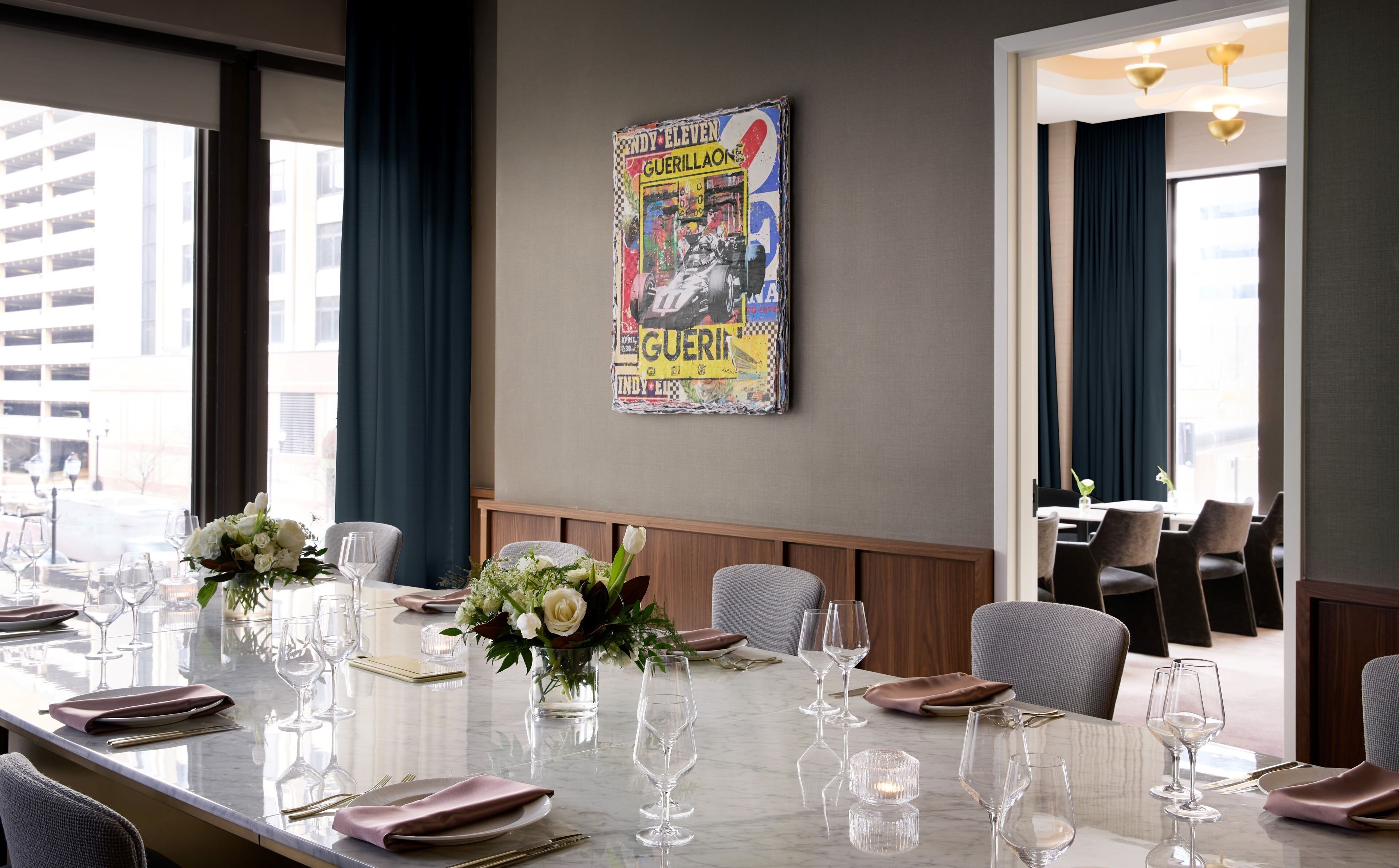 Ein Essbereich mit einem langen Tisch, der für eine Mahlzeit gedeckt ist, einem Poster an der Wand und großen Fenstern mit Vorhängen.