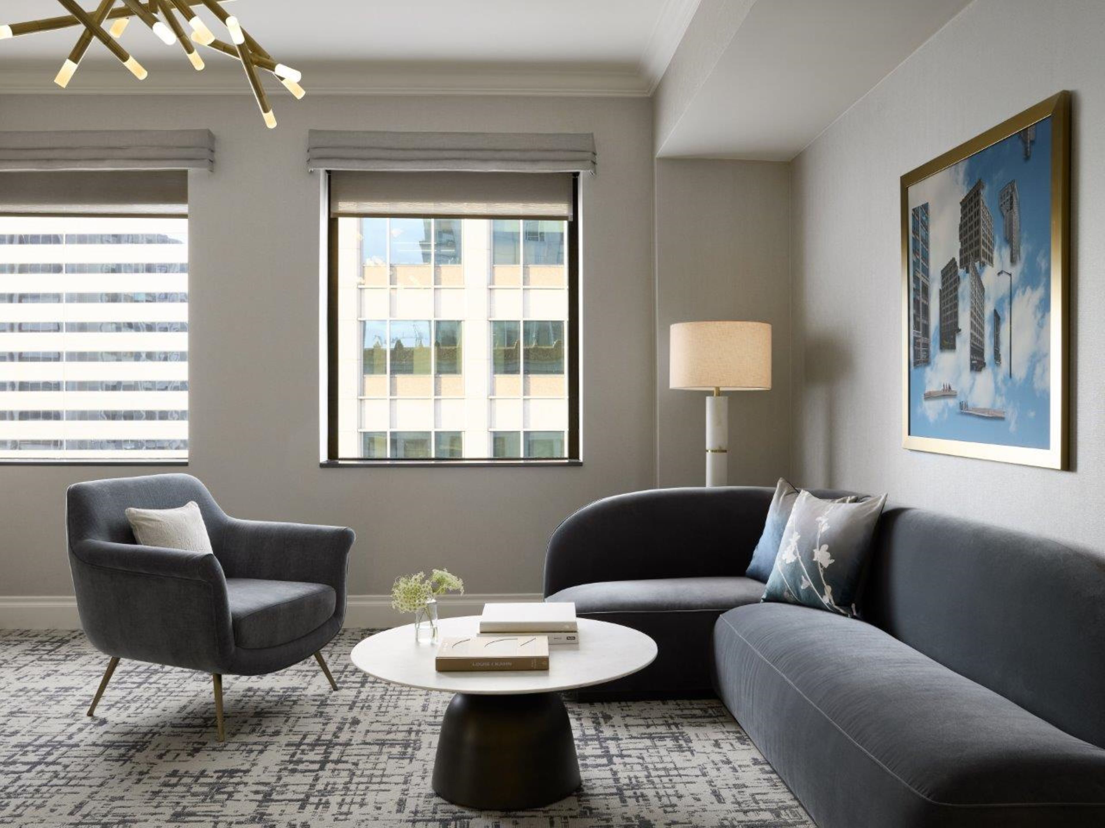A modern living room with a grey sofa, a grey armchair, and a round coffee table. There is a window with blinds, a floor lamp, and a framed painting on the wall.
