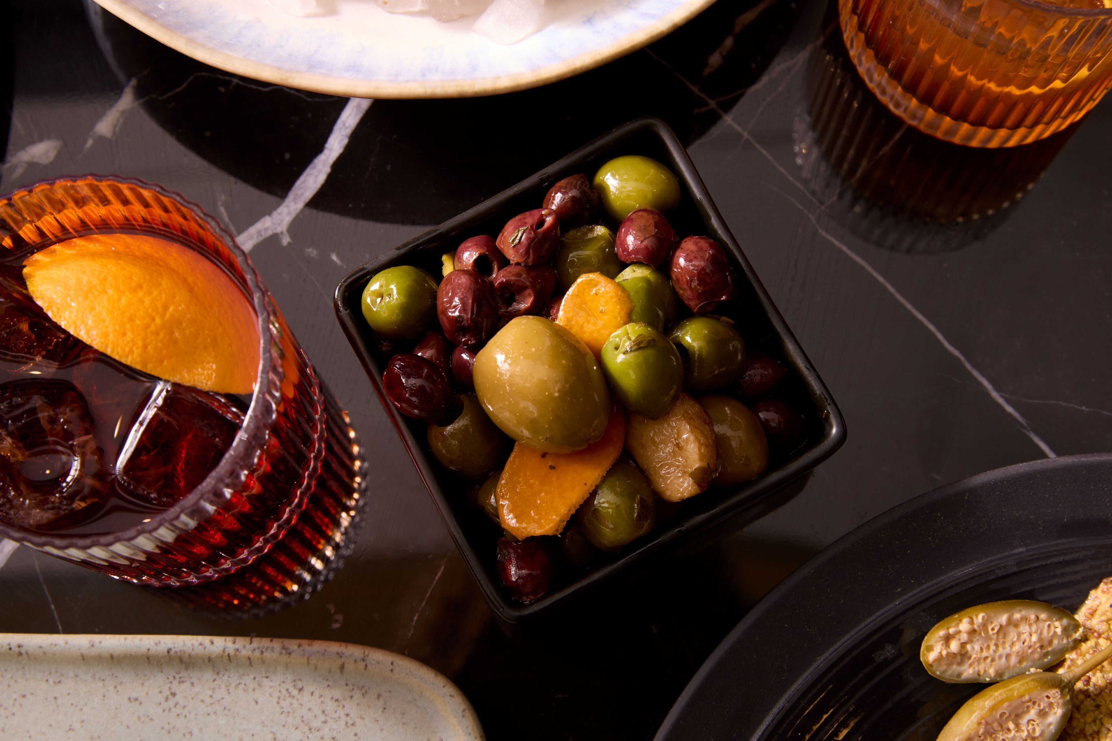 A table with a black tray containing olives, a lemon, and a glass of red beverage.