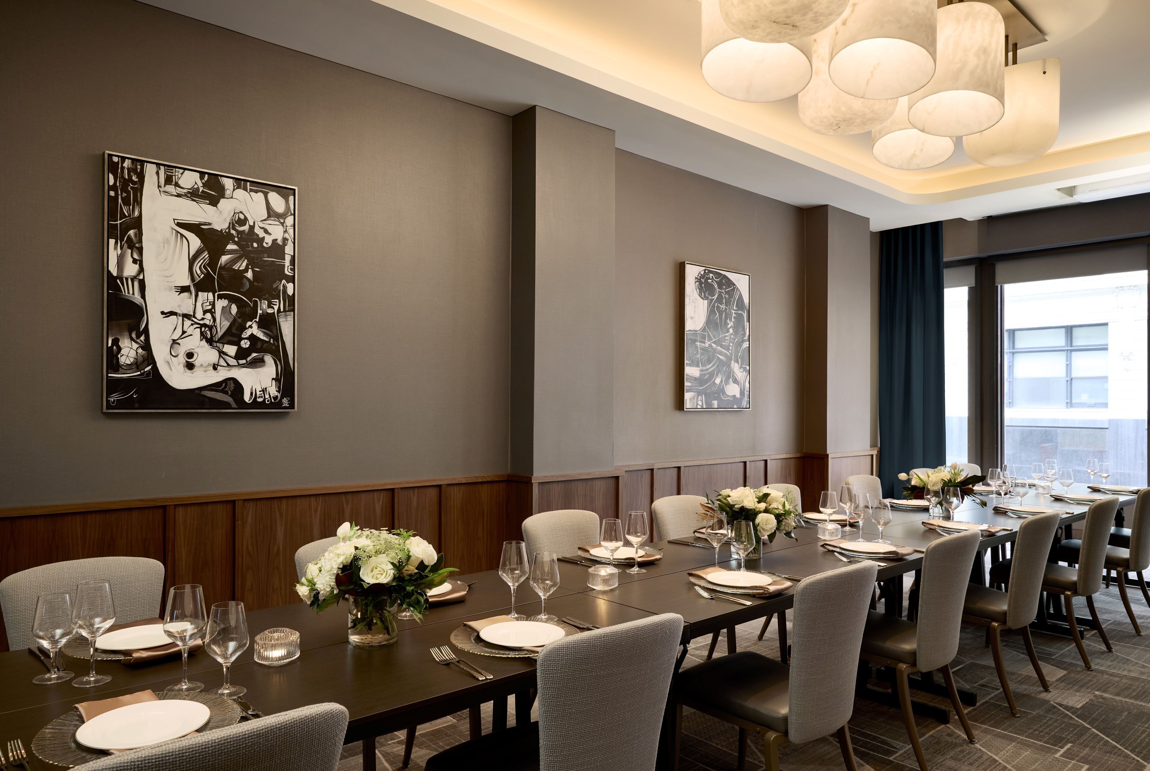 A dining room with a long table set for a meal, flanked by two large black-and-white paintings, and a modern chandelier above.
