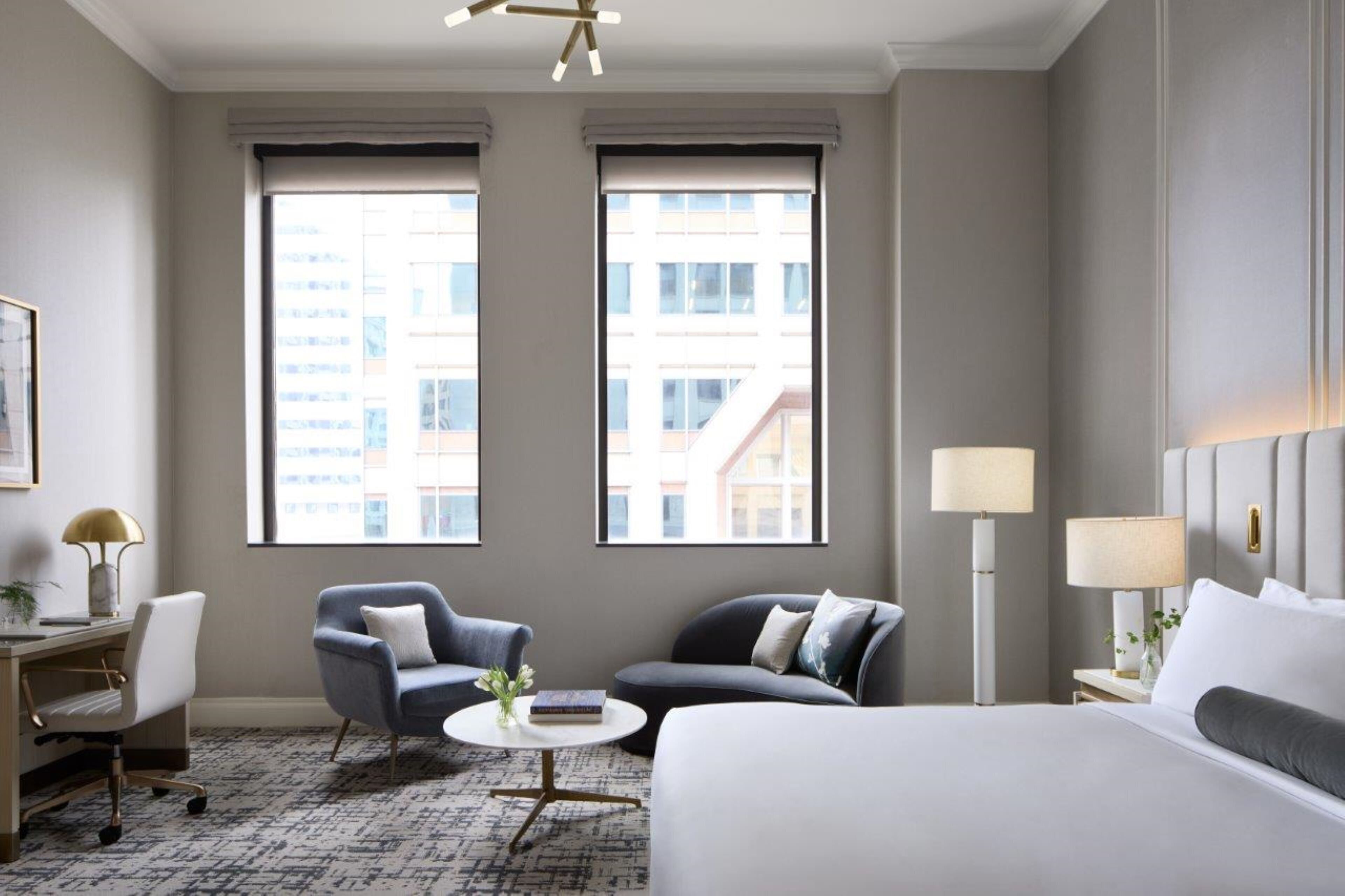 A hotel room with a large bed, two armchairs, a desk, and a small round table. There are two windows providing a view of the cityscape.