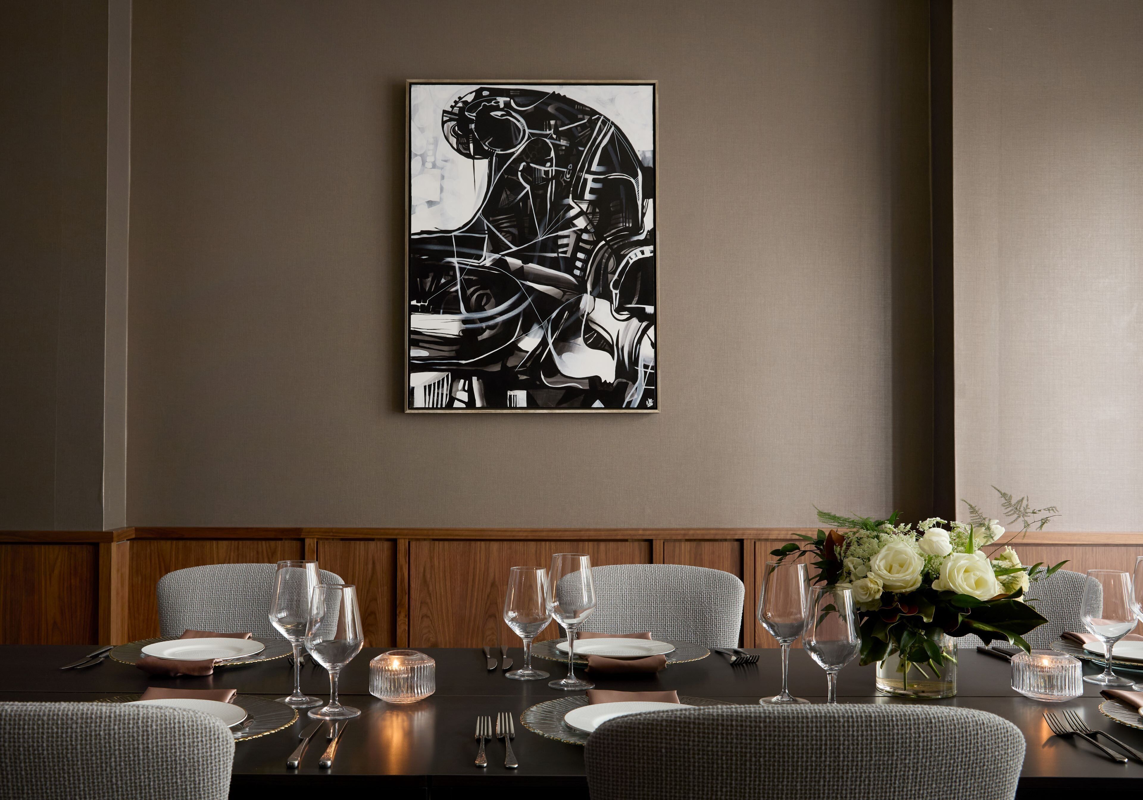 A dining area with a table set for four, including plates, glasses, and cutlery. A vase with flowers is on the table, and a framed artwork is on the wall.