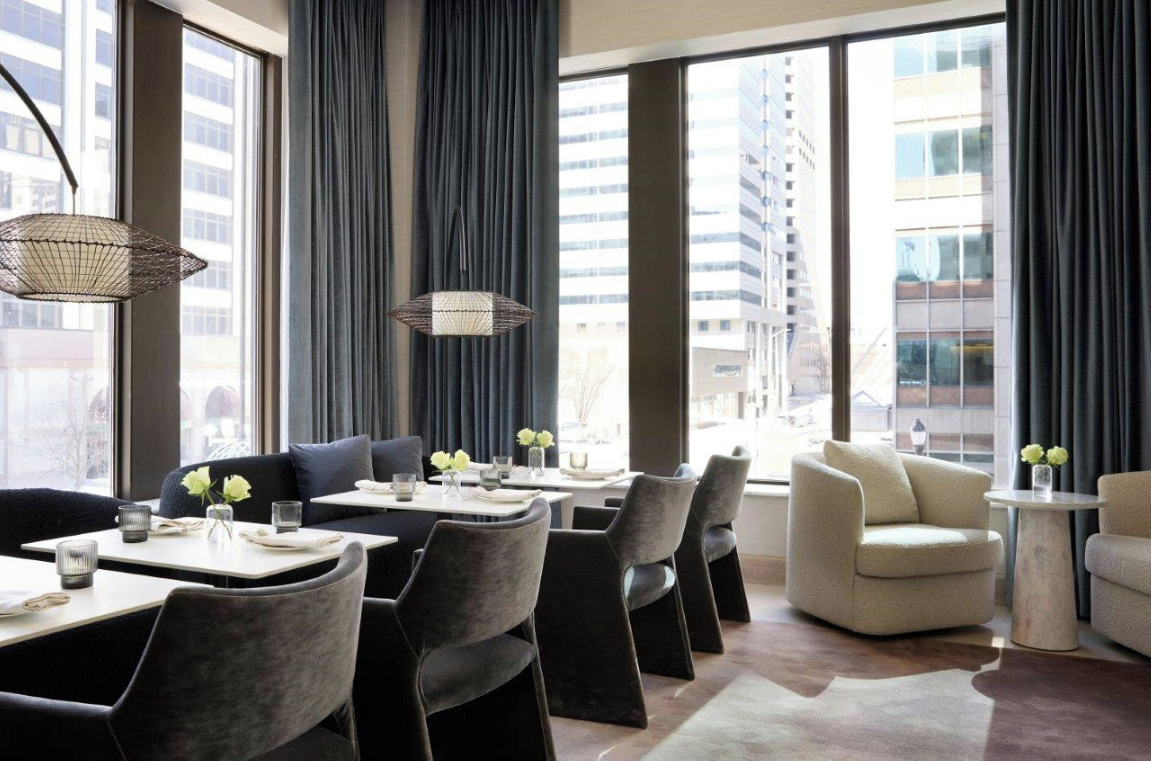 A modern dining area with high tables and chairs, large windows with curtains, and a city view.