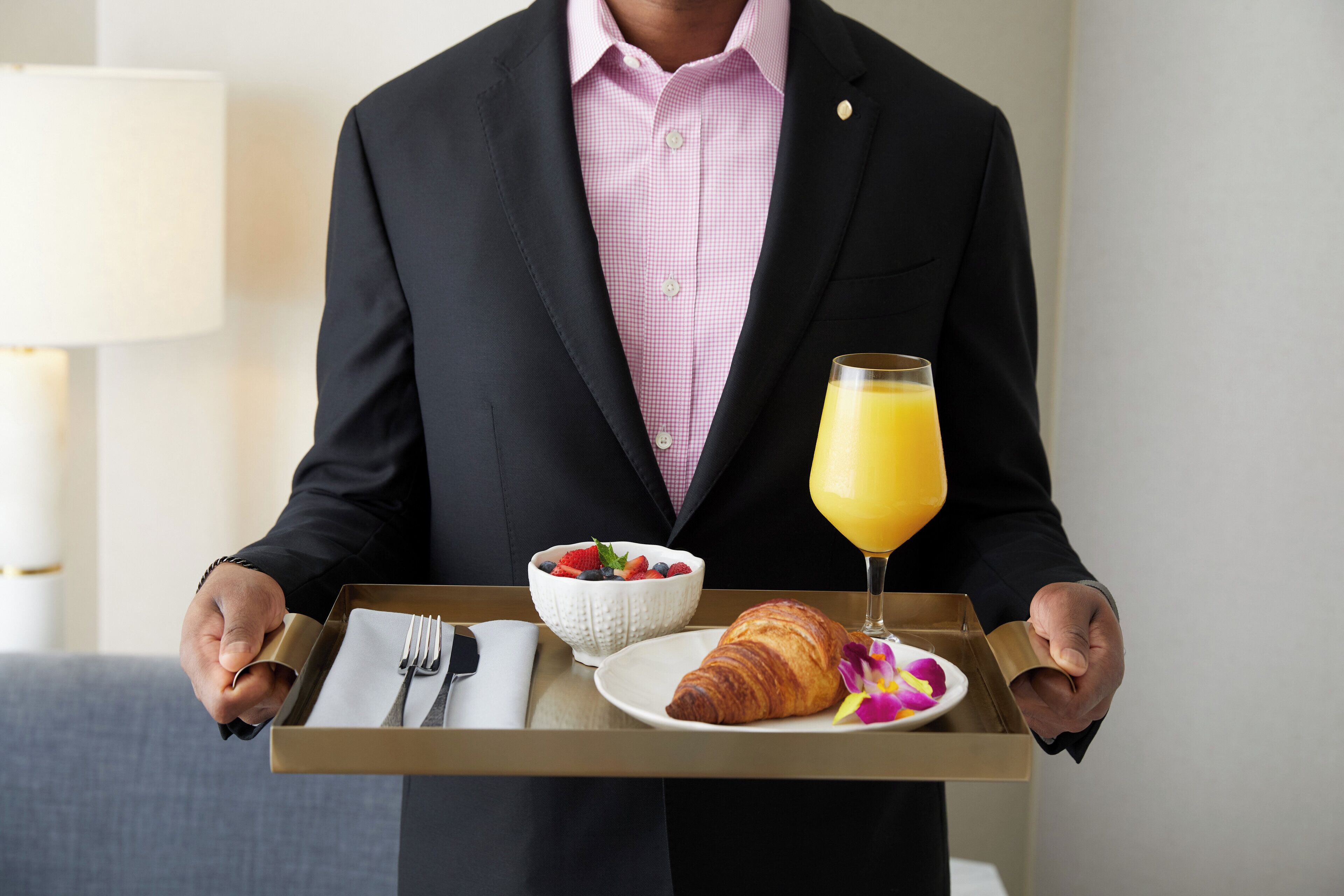 A person in a black suit holding a tray with a croissant, a glass of orange juice, a bowl of fruit, and a napkin.