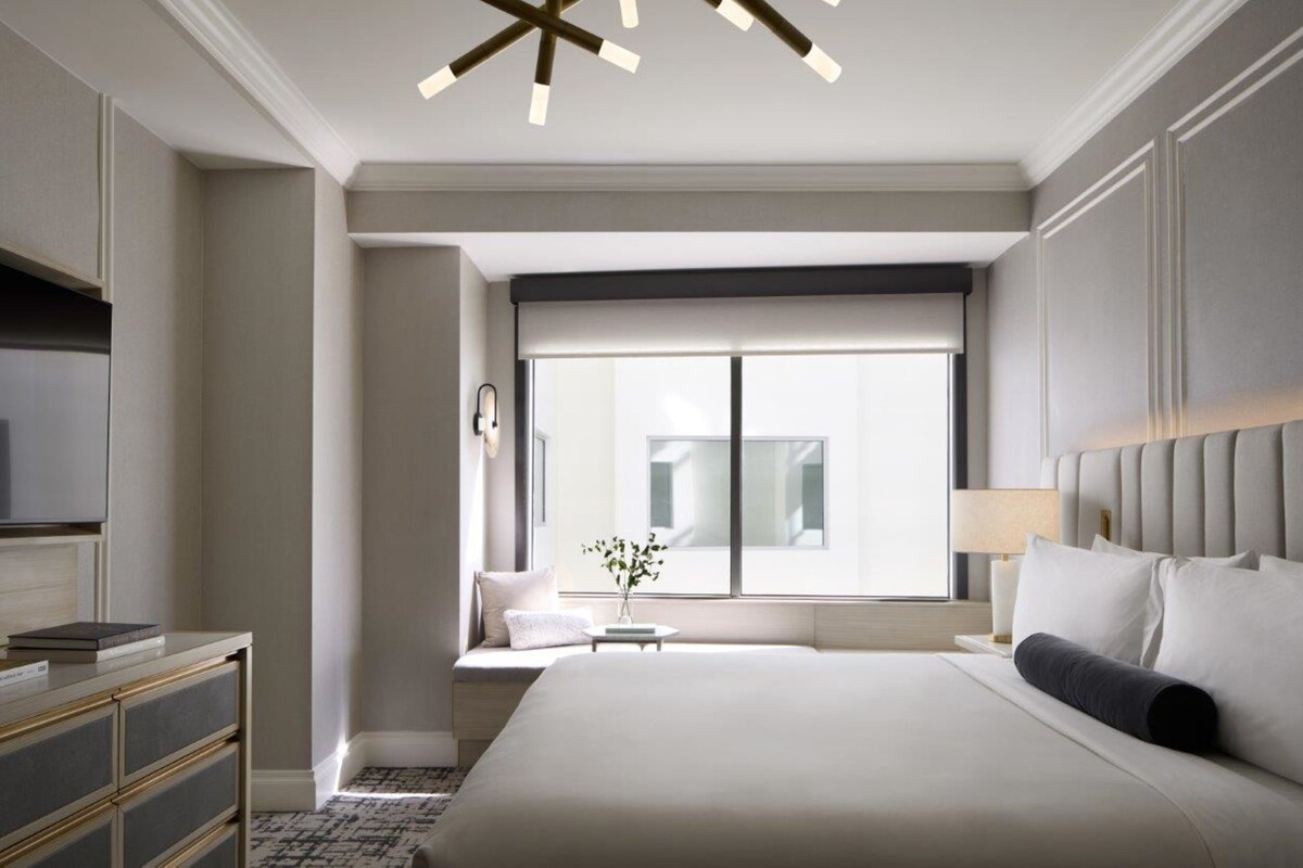 A modern hotel room with a large bed, a television, a small table with a vase, and a window with a view of buildings.