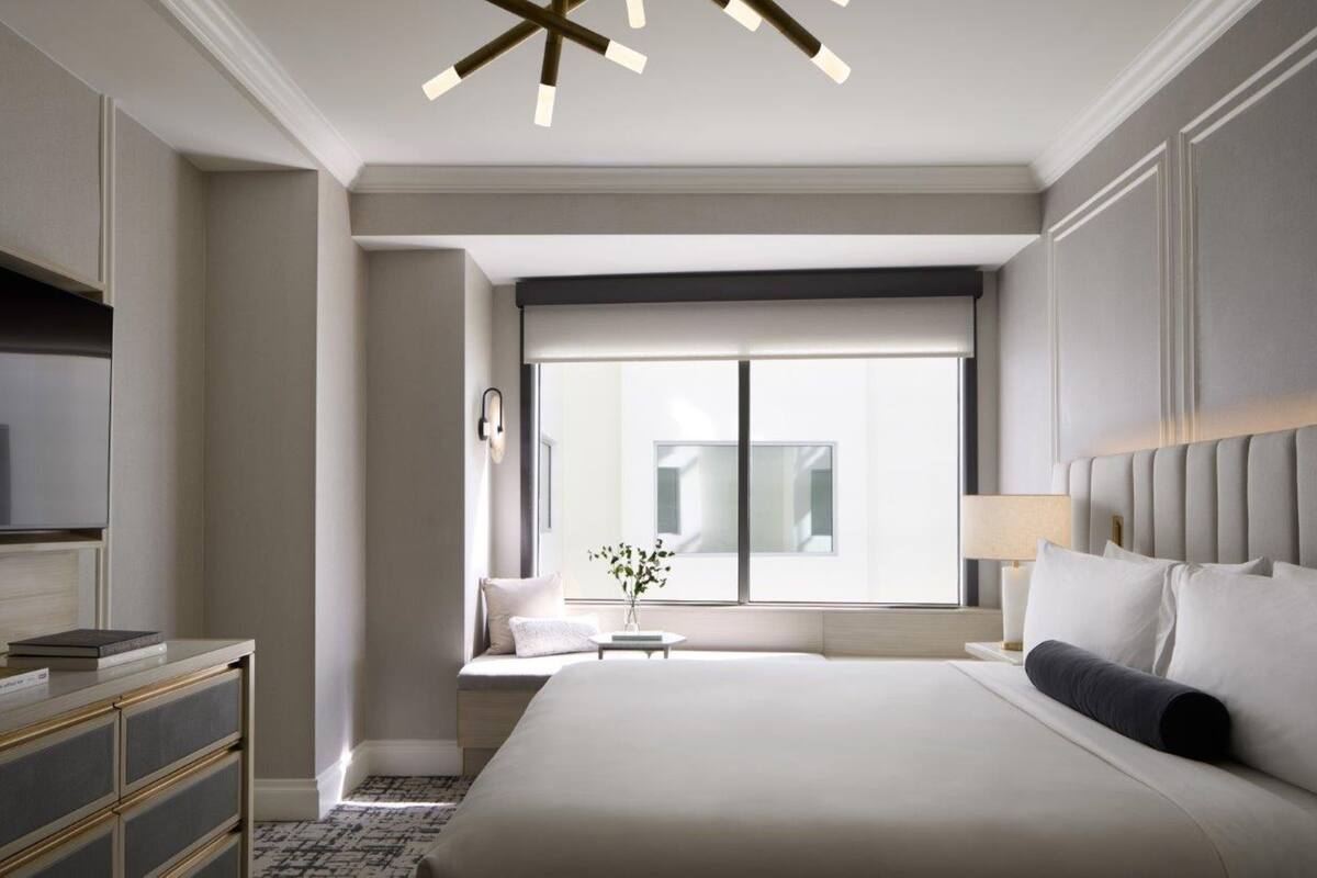 Una habitación de hotel moderna con una cama grande, un televisor, una mesita con un florero y un ventanal con vistas a edificios.