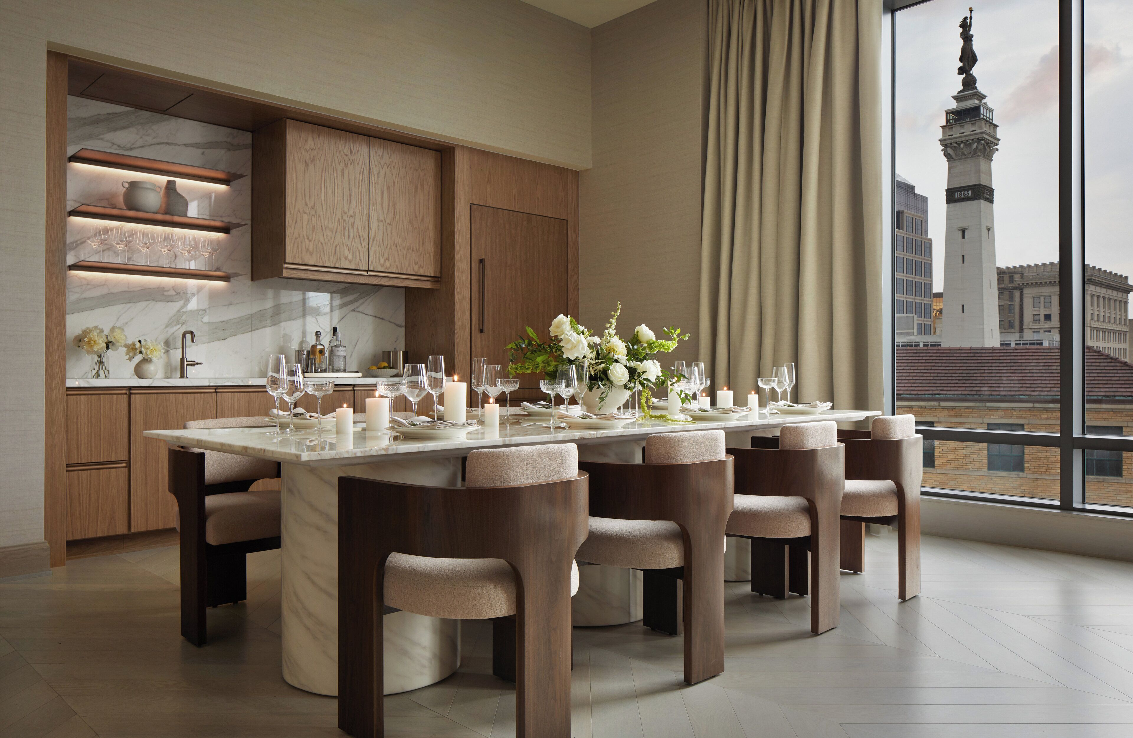 A modern dining area with a large table set for six, wooden cabinetry, and a view of a cityscape with a prominent tower.