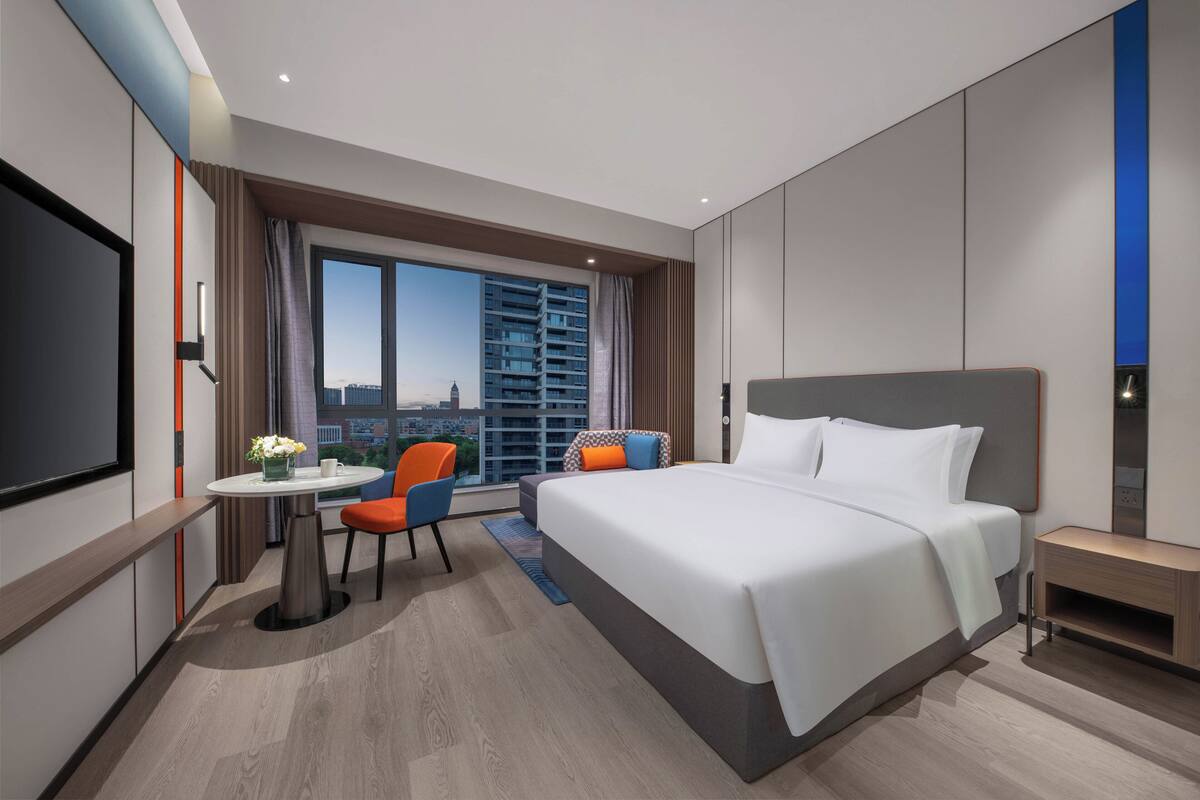 A modern hotel room with a large bed, a desk with a chair, a TV, and a view of the cityscape through large windows.