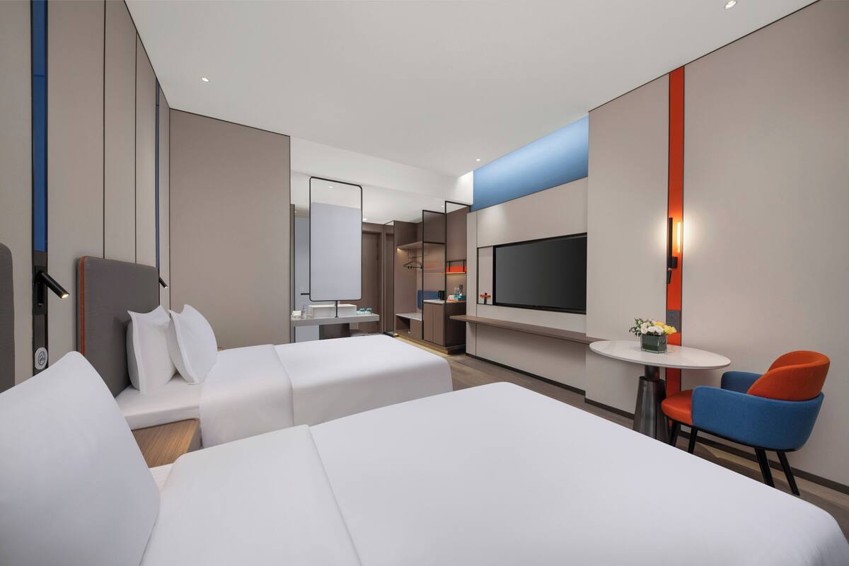 A modern hotel room with two beds, a TV, a small table with a vase, and a chair.
