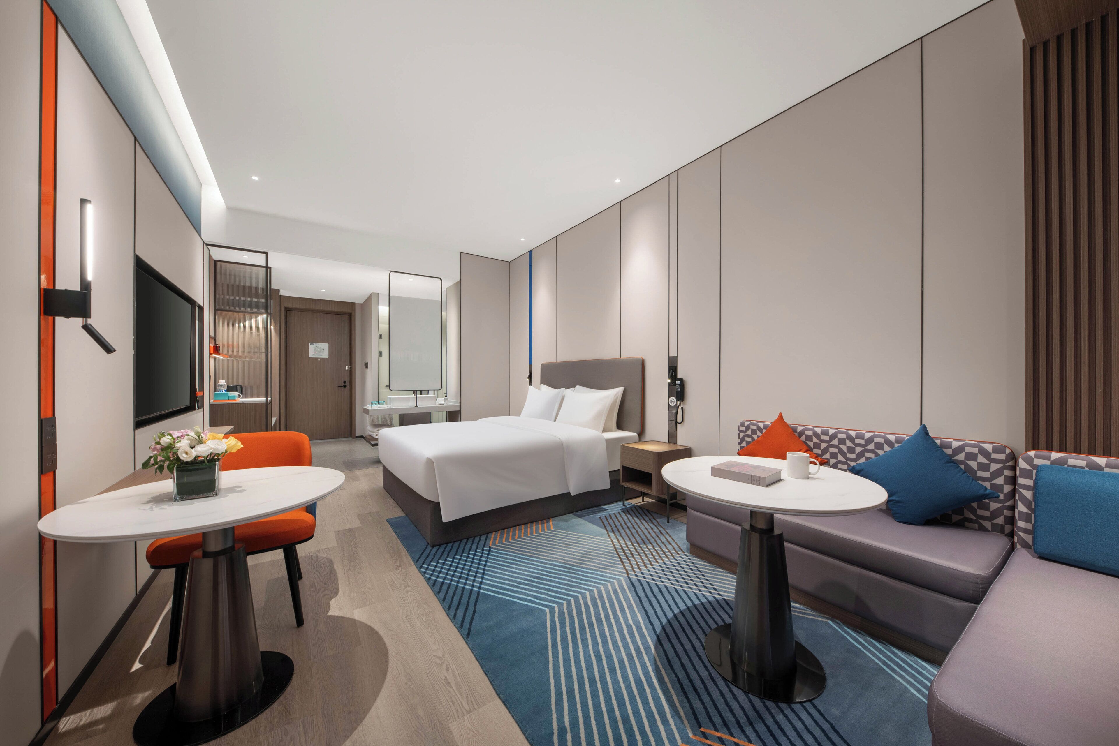 A modern hotel room with a bed, a sofa, a dining table, and a TV.