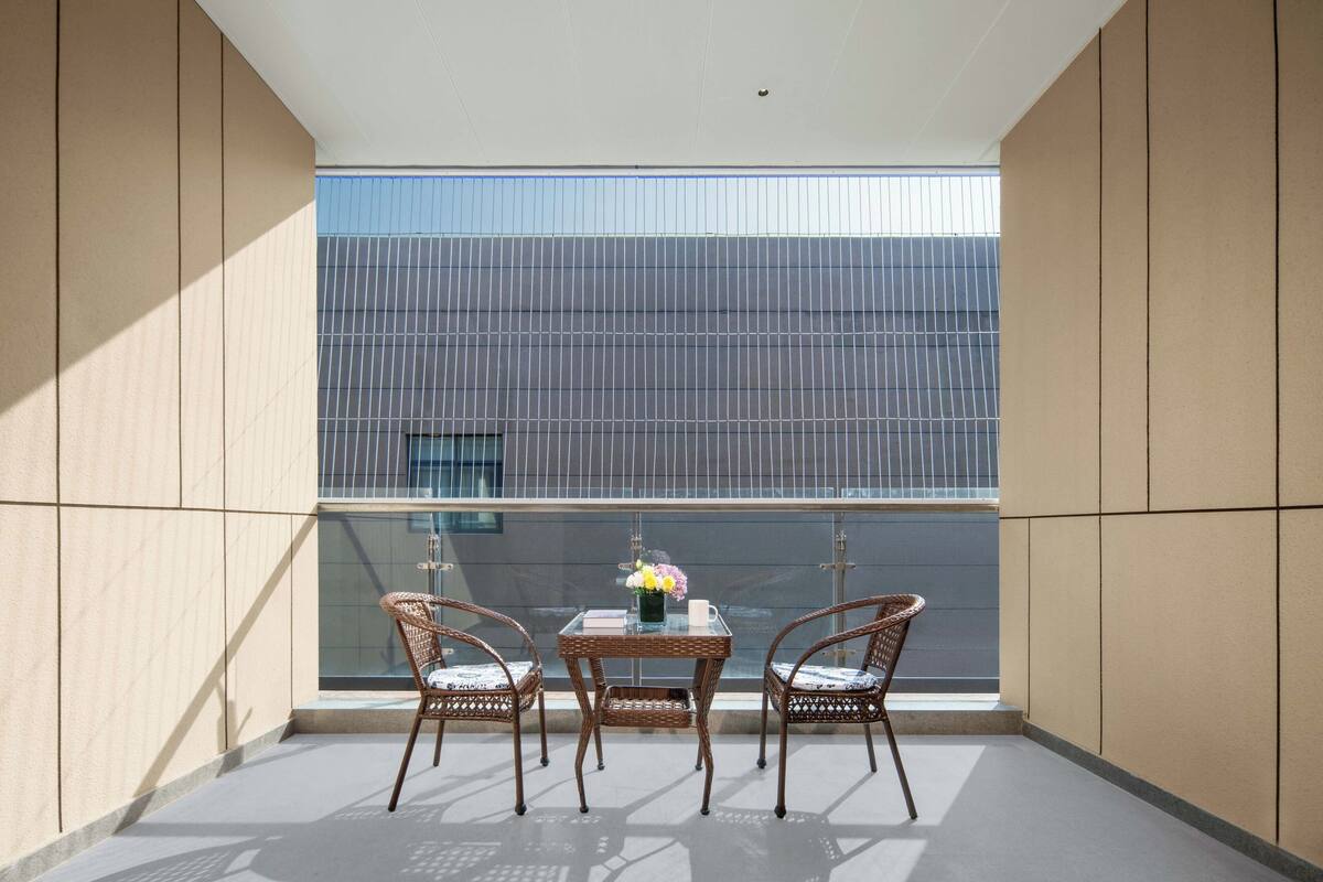 A balcony with a table and two wicker chairs, a vase with flowers, and a view of a building with vertical lines.
