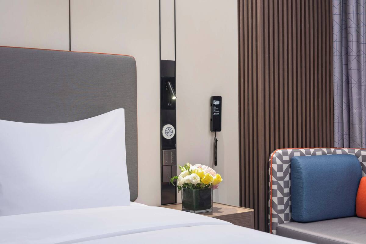 A hotel room with a bed, a small table with a vase of flowers, a sofa with cushions, a telephone, and a wall-mounted control panel.