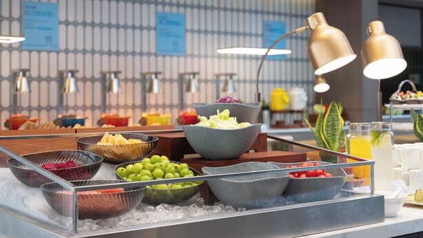 Un buffet de petit-déjeuner proposant une variété de fruits et de boissons, un panneau indiquant « DÉBUT DU PETIT-DÉJEUNER » et un aménagement intérieur moderne.