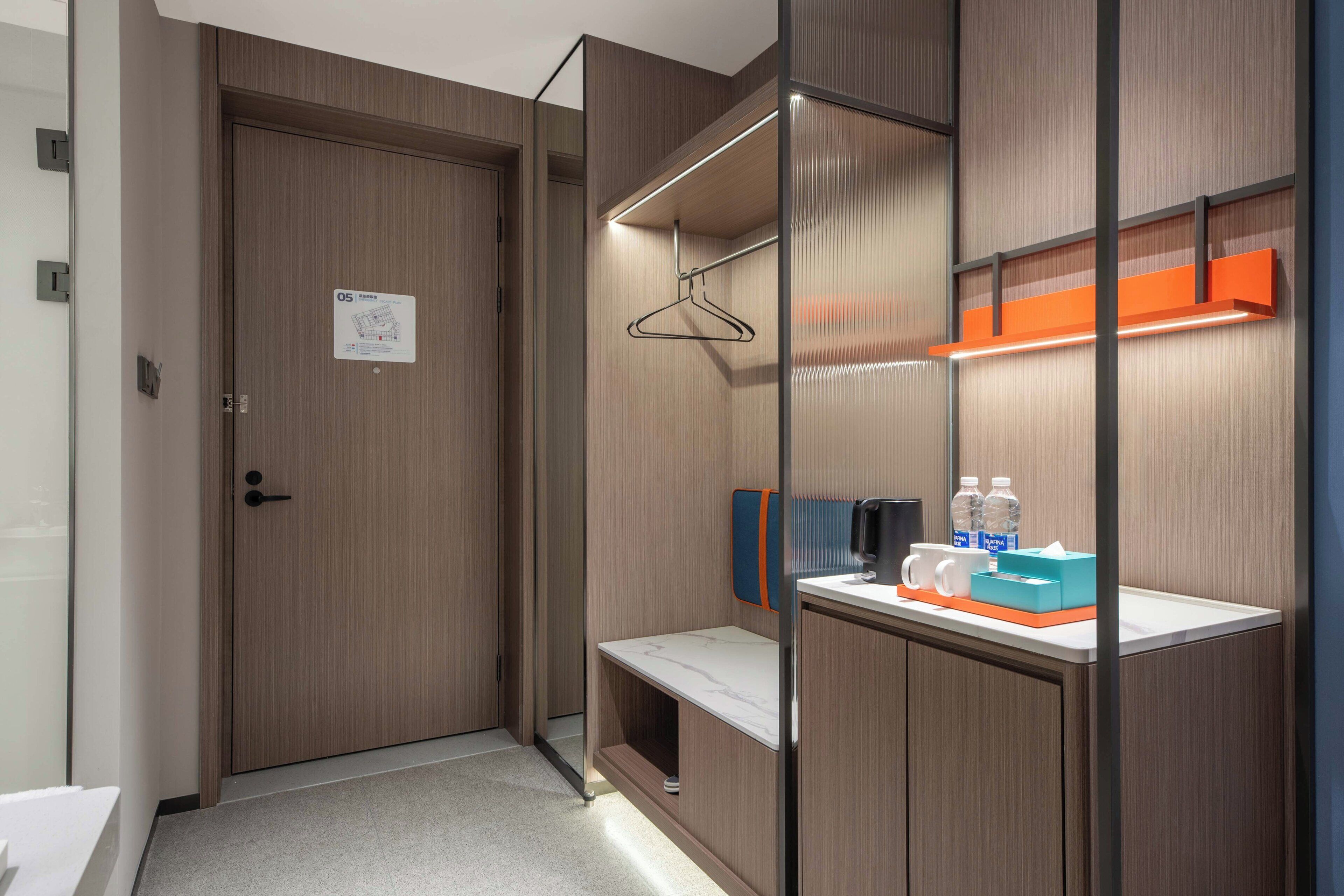 A modern hotel room with a wooden door, a small bench, a mirror, and a shelf with toiletries.