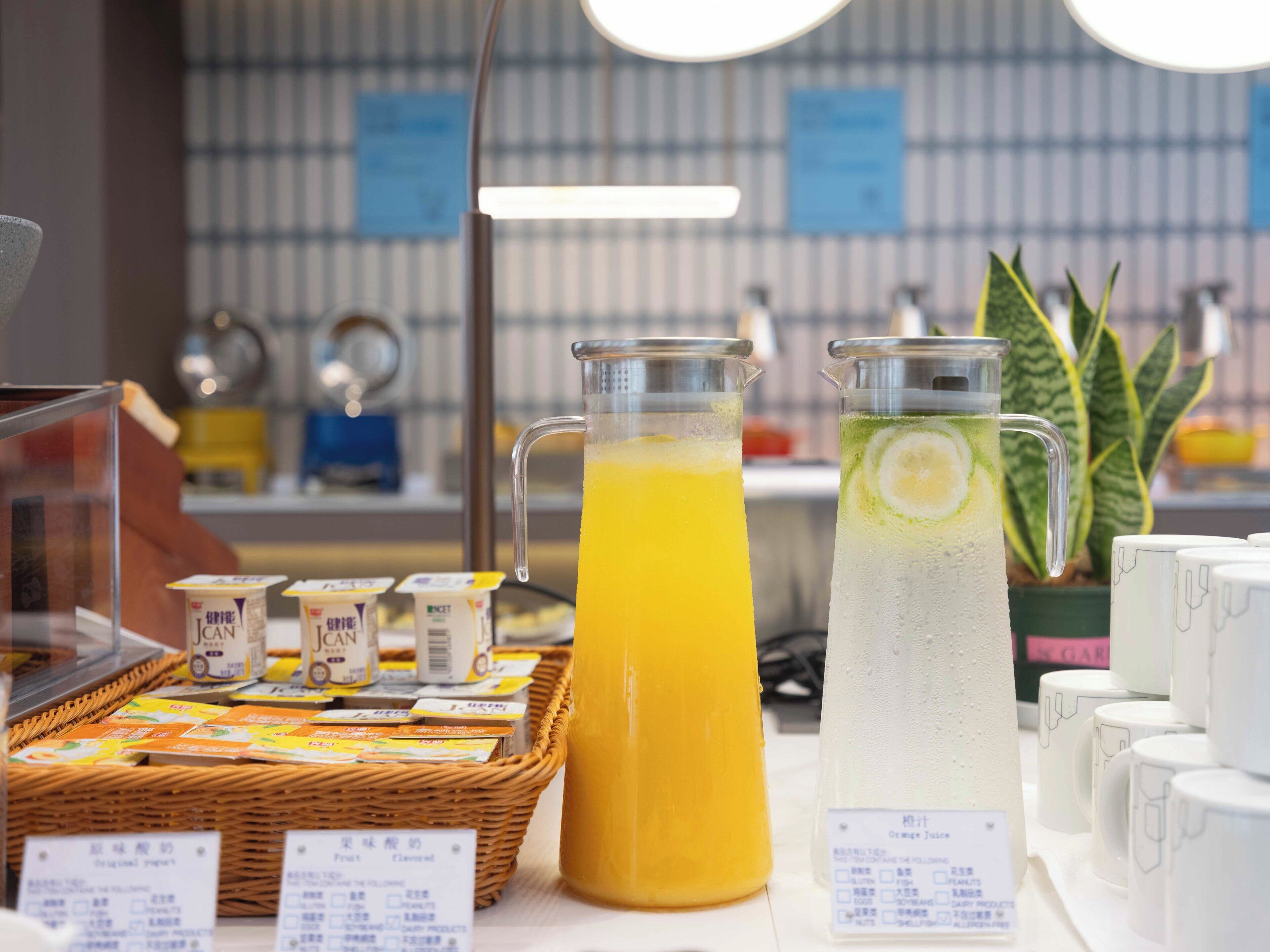 A beverage stand with two pitchers, one with orange juice and the other with a lime-infused drink, and a basket of packaged snacks.