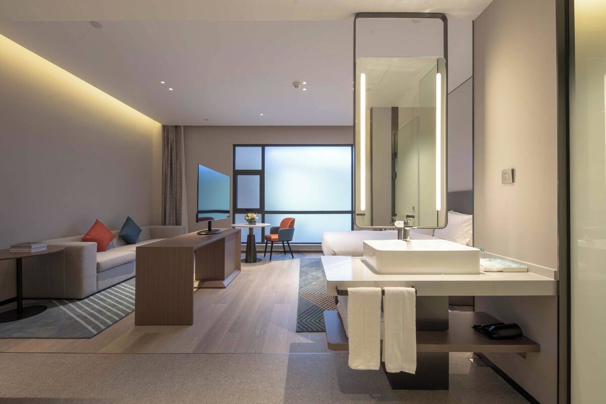 A modern hotel room with a large bed, a sitting area with a sofa and armchairs, a dining table, and a large mirror.