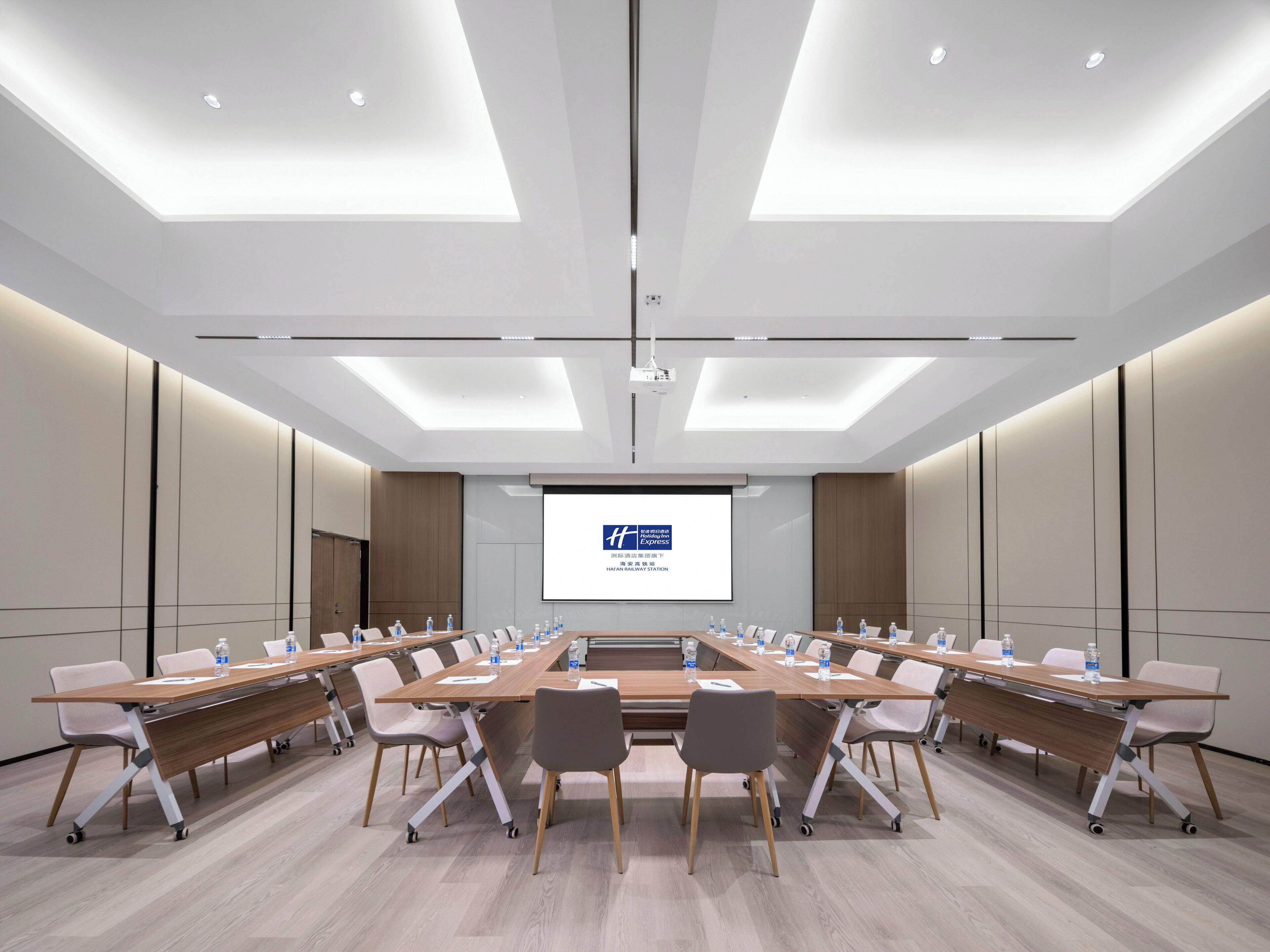 A conference room with a long table, chairs, a projector screen, and a ceiling with recessed lighting.