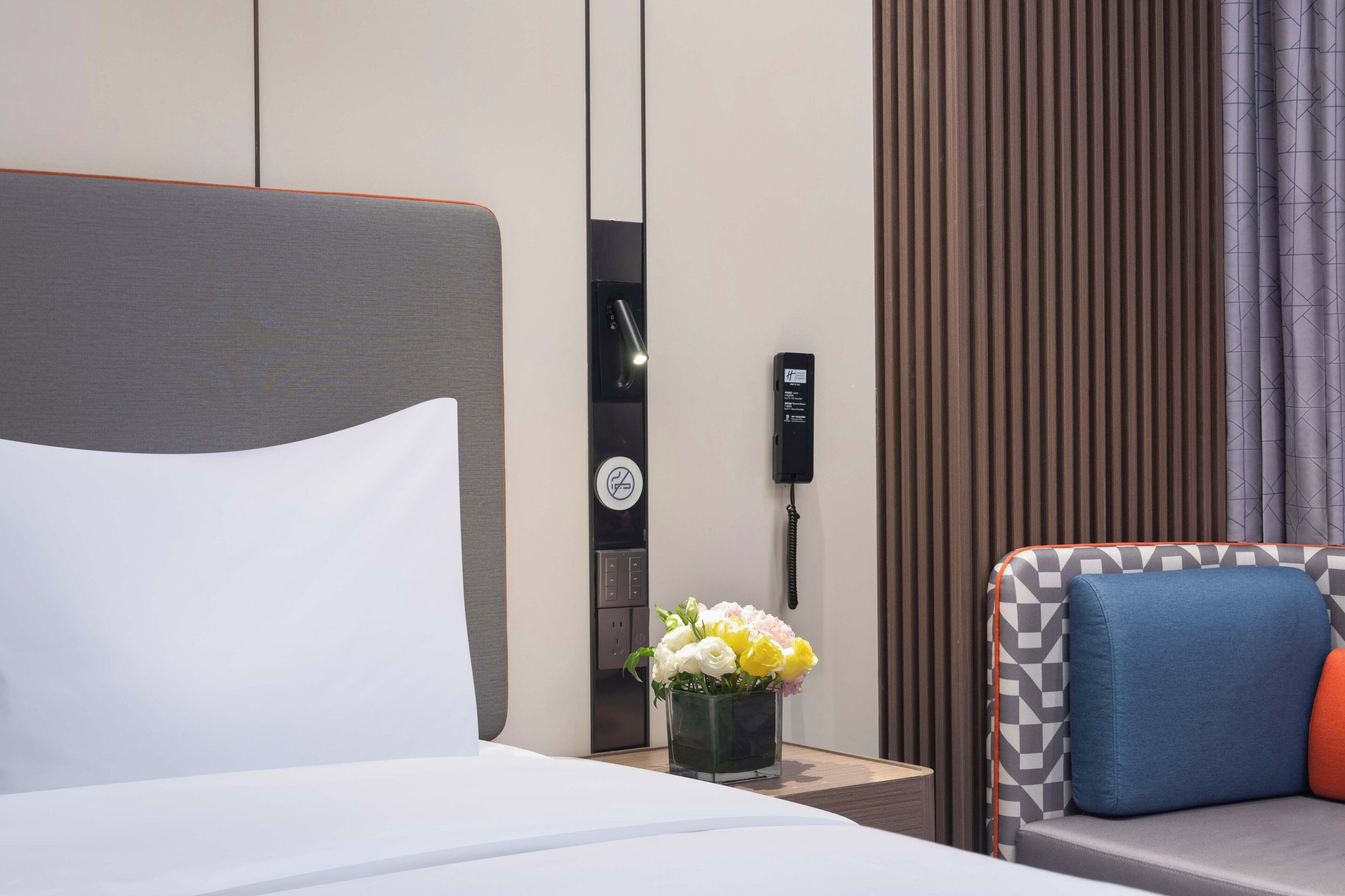 Habitación de hotel con cama, una pequeña mesa con un jarrón de flores, un sofá con cojines, un teléfono y un panel de control en la pared.