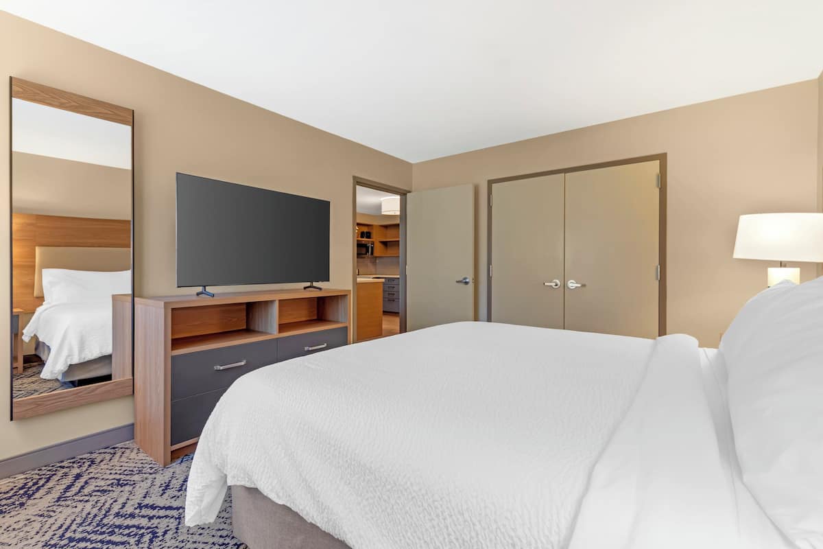 A hotel room with a large bed, a flat-screen TV on a wooden stand, a wardrobe, a mirror, a door, and a lamp.