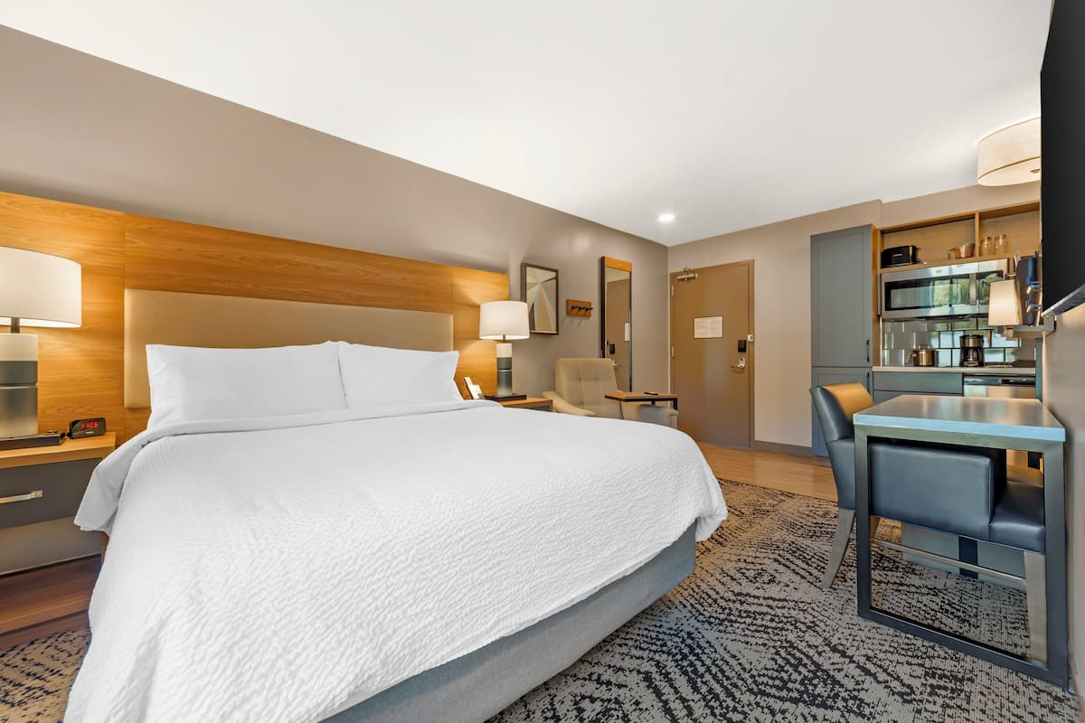 A modern hotel room with a large bed, bedside tables, a desk, and a kitchenette.