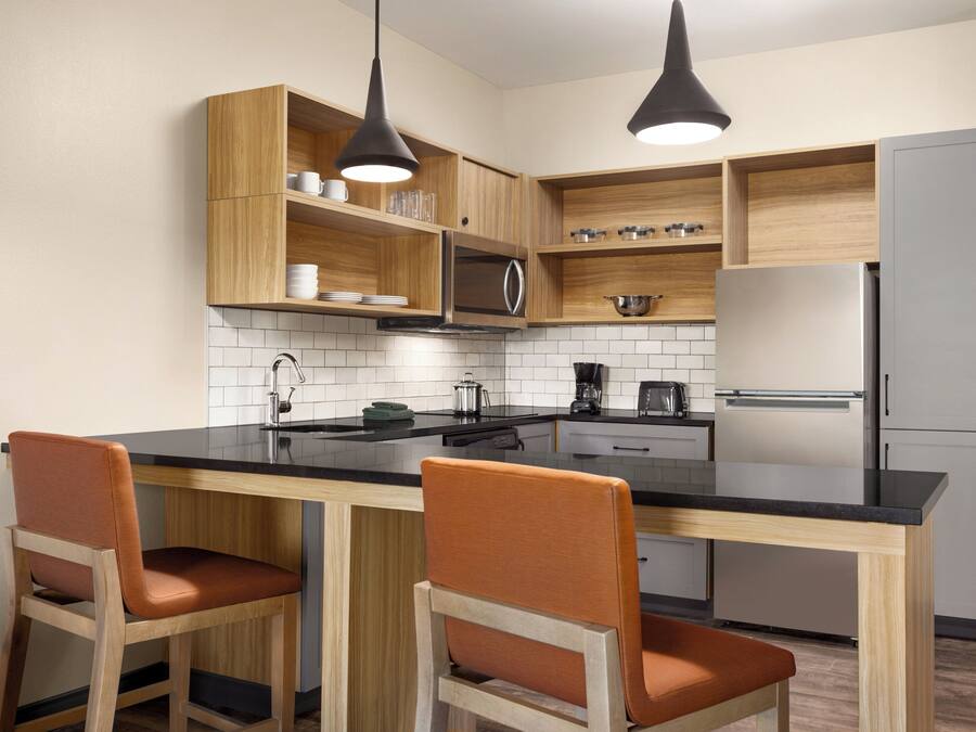 A modern kitchen with wooden cabinets, a black countertop, two orange chairs, and a stainless steel refrigerator.