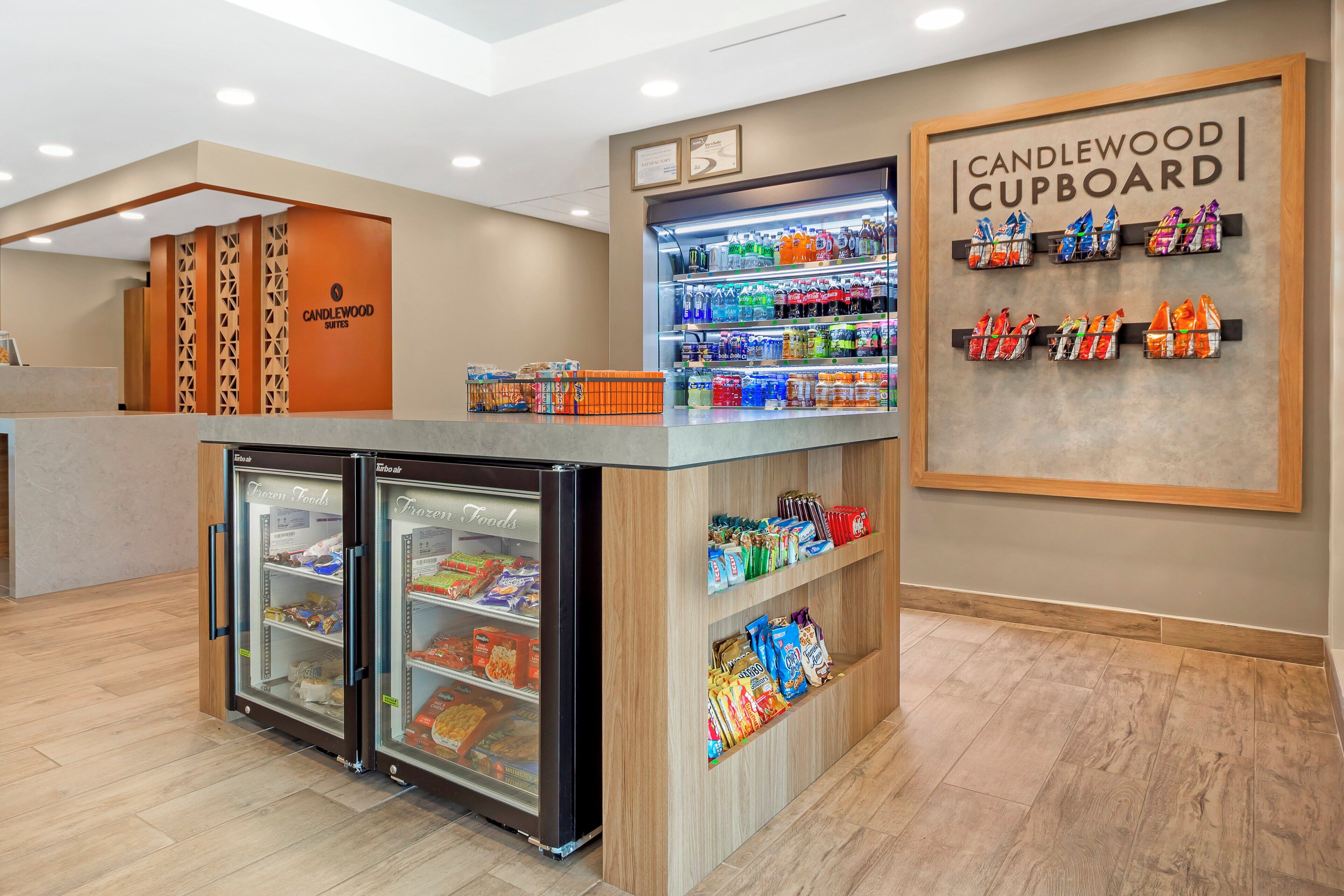 A hotel reception area with a refrigerated display case, a freezer, and a wall-mounted sign for the Candlewood Cupboard.