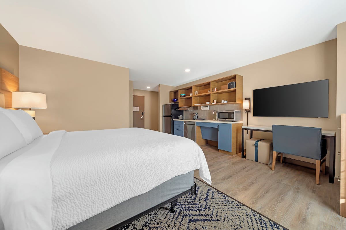A hotel room with a bed, a desk, a chair, a TV, and a kitchenette.