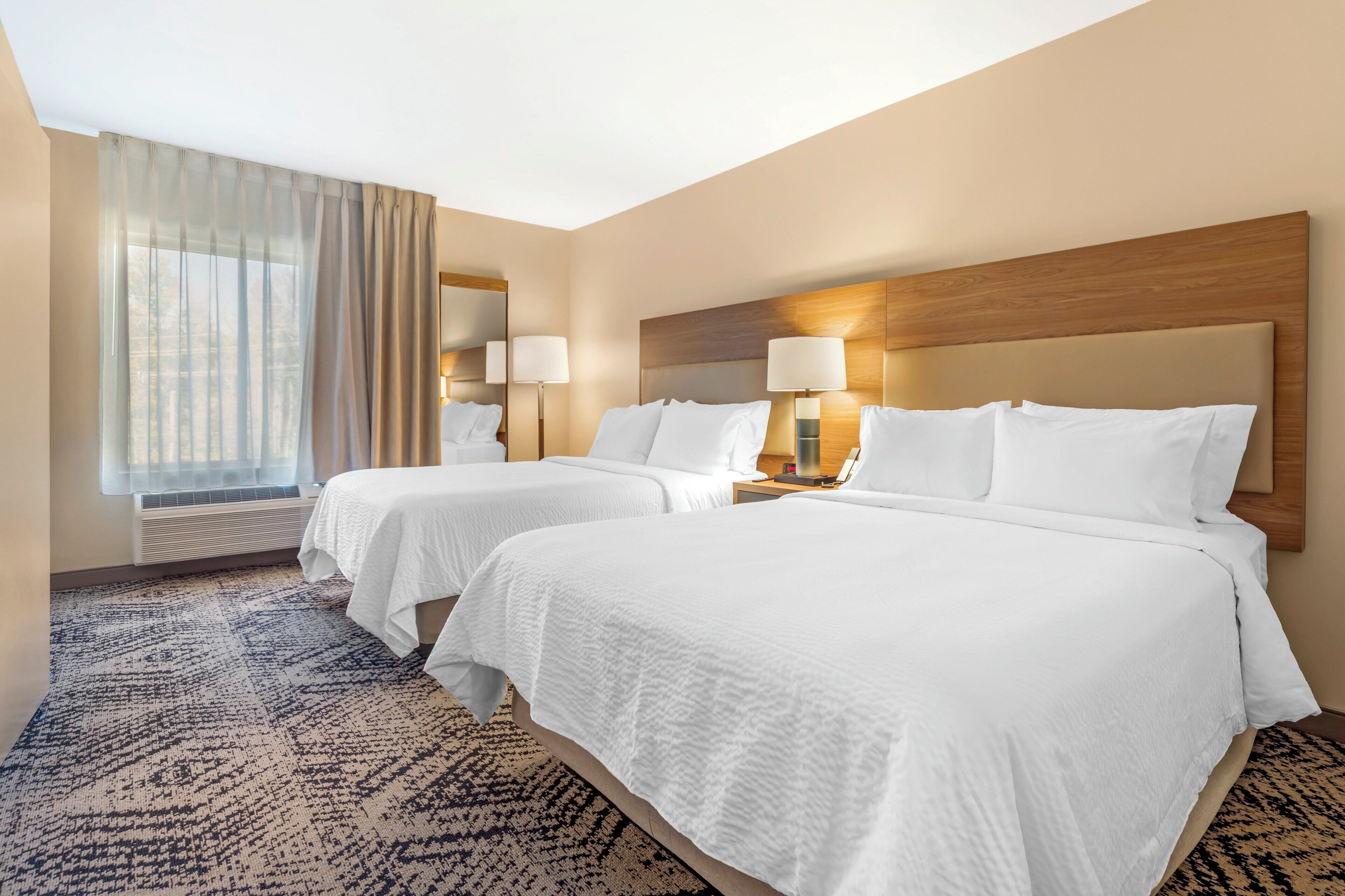 Une chambre d’hôtel avec deux lits, une grande fenêtre avec des rideaux, une moquette à motifs et une tête de lit en bois.
