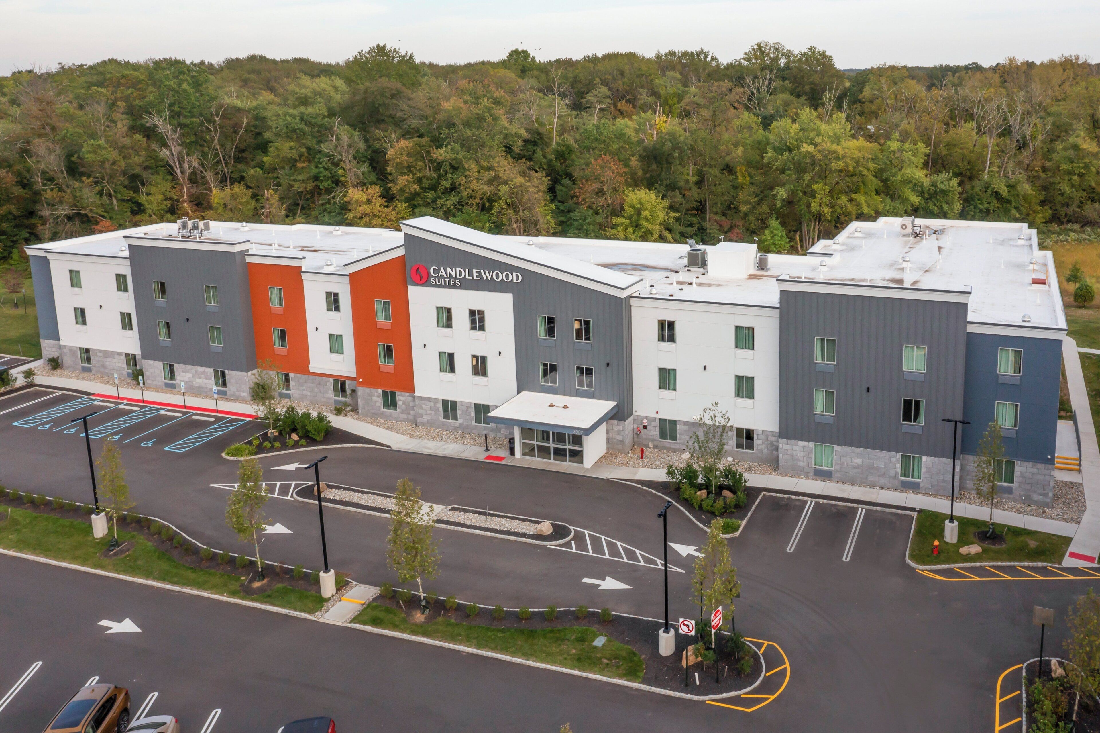 A multi-story hotel with a modern design, featuring a mix of white, grey, and orange facades, and a parking lot with marked spaces and directional arrows.