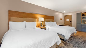 A hotel room with two beds, a wooden headboard, a nightstand with a lamp, a desk, and a kitchenette.
