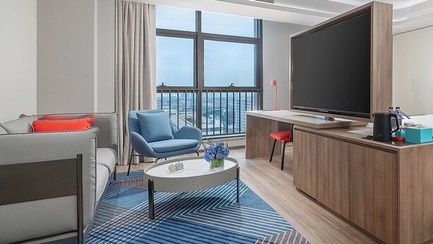 A modern hotel room with a sofa, a blue armchair, a small round table with a vase of flowers, a large TV, and a wooden cabinet. - Holiday Inn Express Foshan Shunde Lecong (Foshan)