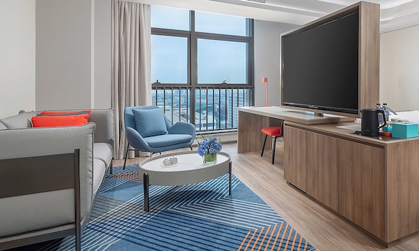 Ein modernes Hotelzimmer mit einer Couch, einem blauen Sessel, einem kleinen runden Tisch mit Blumenvase, einem großen Fernseher und einem Holzschrank.