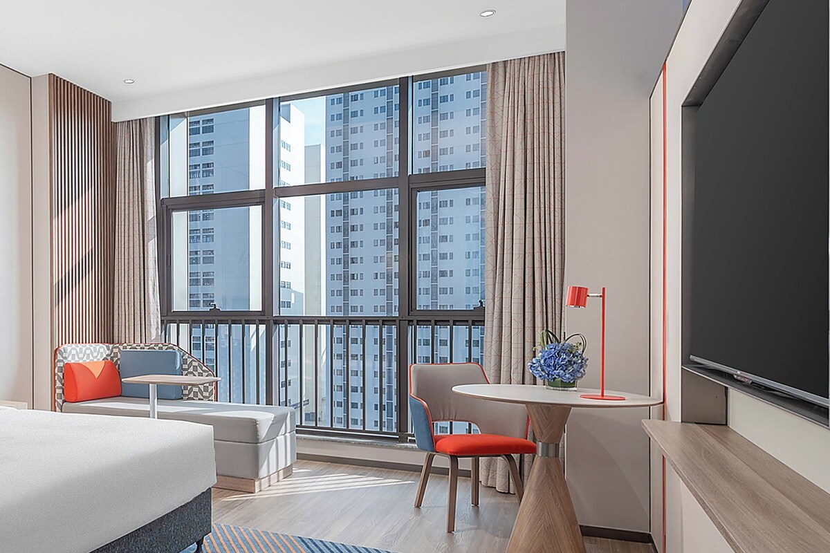 A modern hotel room with a large window, a bed, a desk, and a chair.