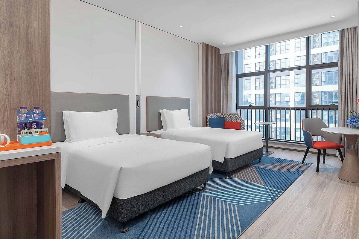 A hotel room with two beds, a desk with a chair, a small table with a lamp, and a large window offering a city view.