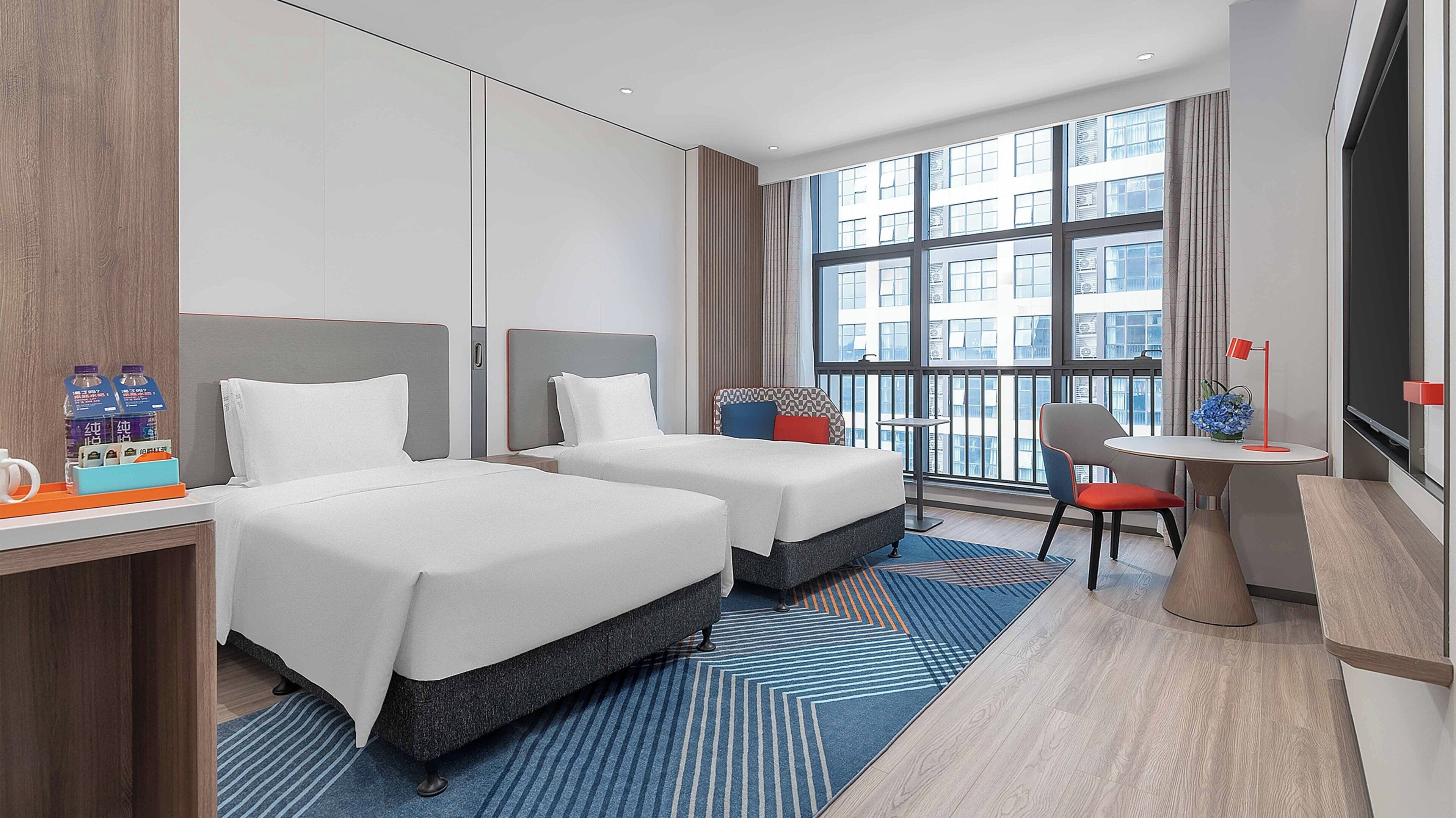 A hotel room with two beds, a desk with a chair, a small table with a lamp, and a large window offering a city view.