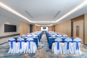 A conference room with rows of tables and chairs, a large screen at the front, and a blue carpet.