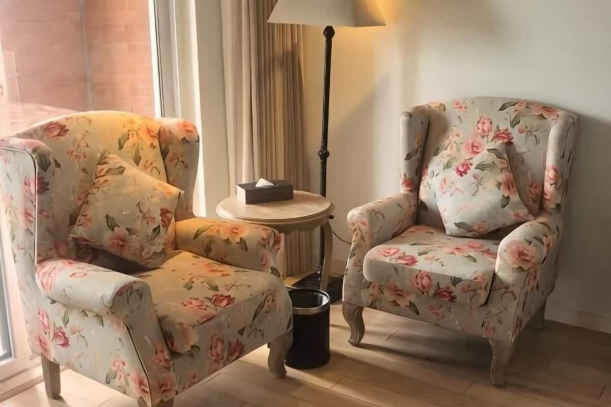 A room with two floral armchairs, a small round table with a lamp, and a trash bin.