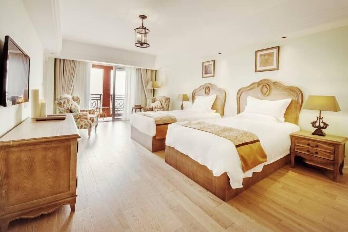 A hotel room with two beds, a wooden dresser, a desk, and a balcony with curtains.