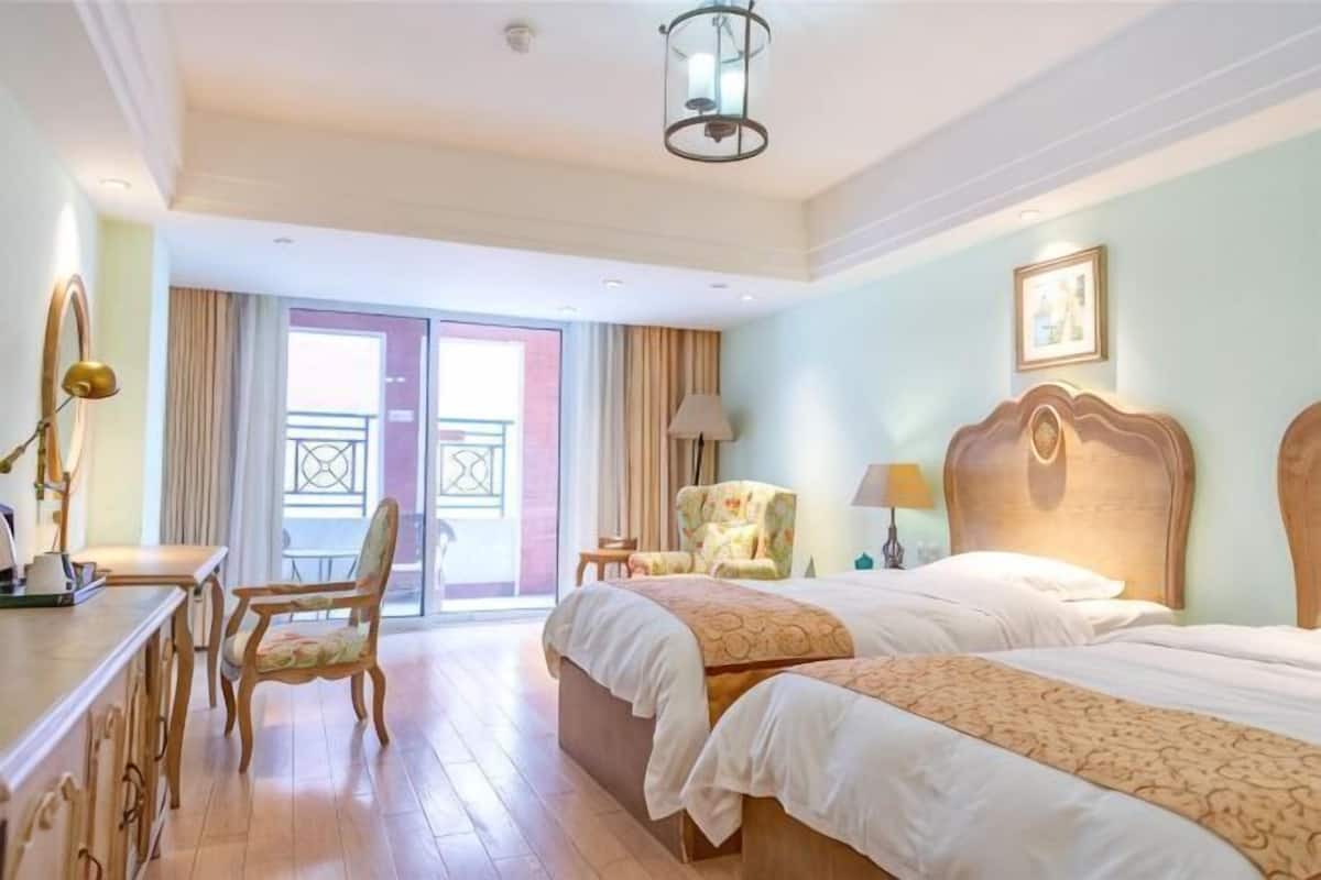 A hotel room with two beds, a desk, a chair, a balcony, and a lamp.