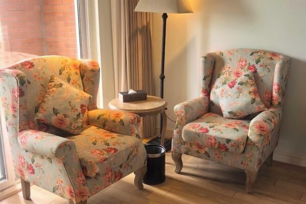 Two floral armchairs, a lamp, and a small table with a tissue box in a room with a brick wall and a window.