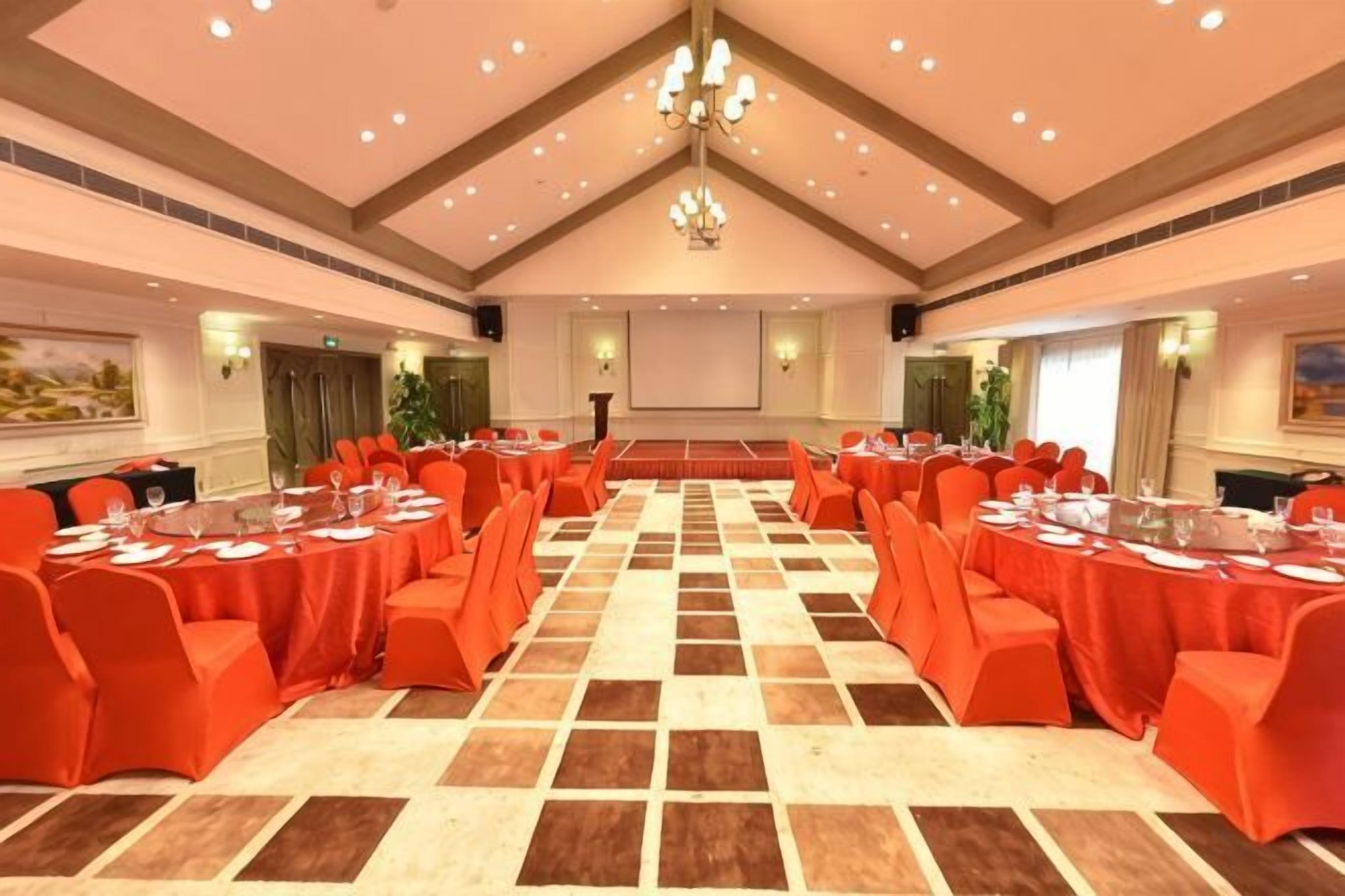 Une salle de conférence avec un plafond haut, un lustre et une scène à l’avant. Des tables sont disposées, recouvertes de nappes rouges, et des chaises sont placées autour.