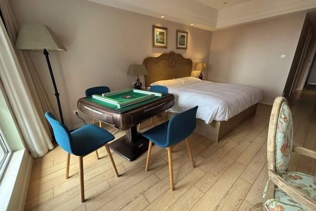 A hotel room with a bed, a desk with a green board game, two chairs, a lamp, and two framed pictures on the wall.