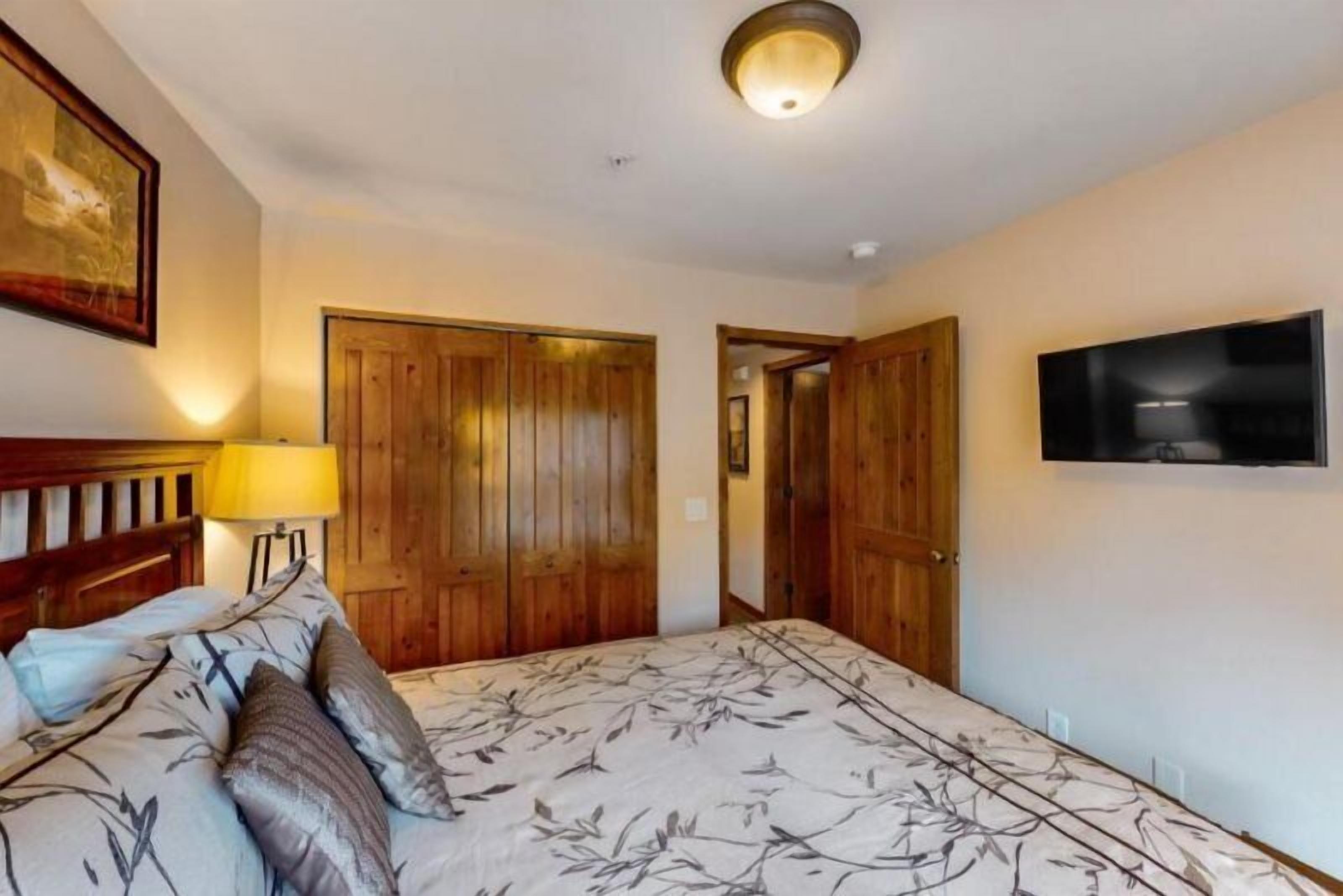 Un dormitorio con cama, armario de madera, televisor montado en la pared y una lámpara.