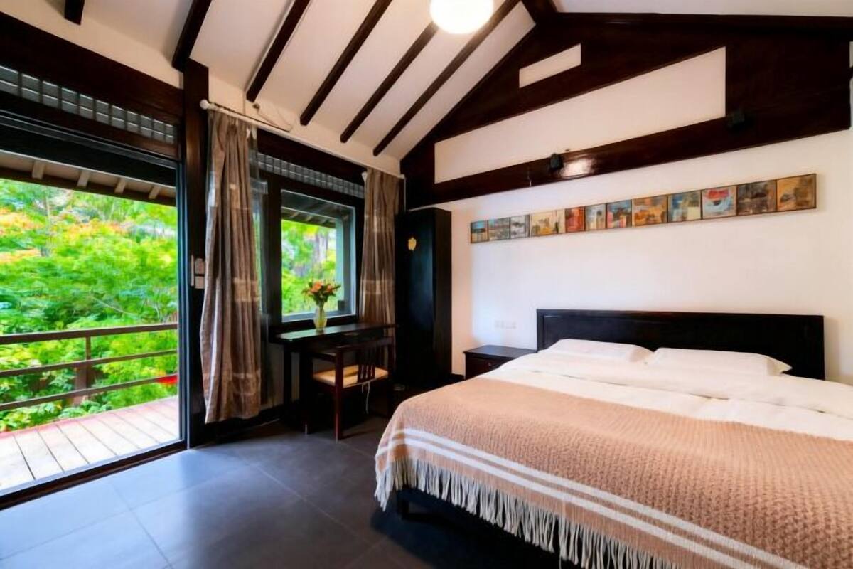 A bedroom with a large bed, a desk, and a window with a view of greenery.
