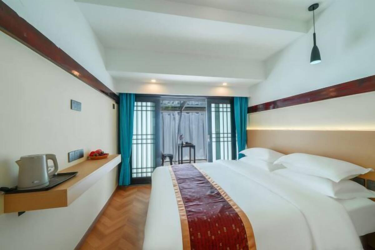 A hotel room with a bed, a bedside table with a kettle, a small table with a chair, and a window with curtains.