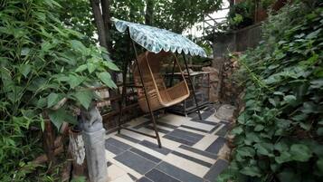 Un jardín con una silla mecedora bajo un dosel azul, rodeado de follaje y un piso de baldosas.