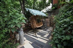 Un jardín con una silla mecedora bajo un dosel azul, rodeado de follaje y un piso de baldosas.