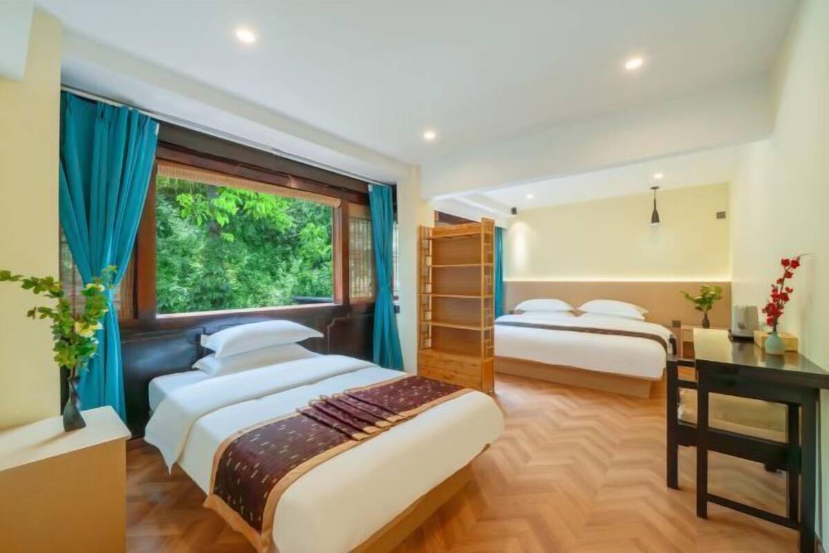 A hotel room with two beds, a desk, and a view of greenery outside.