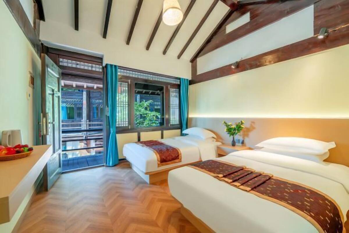 A hotel room with two beds, a wooden ceiling, a view through a glass door, and a small table with fruit.