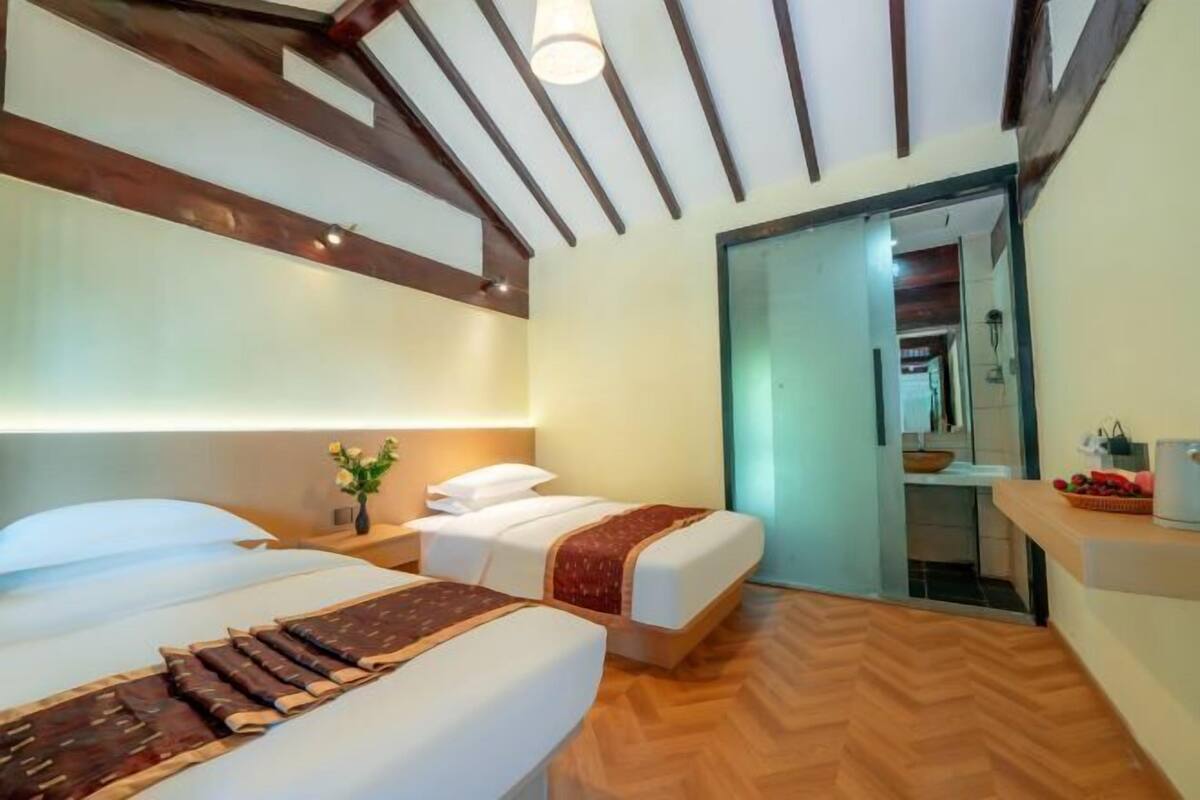 A hotel room with two beds, a wooden ceiling, a small table with a vase of flowers, and a view into the hallway.